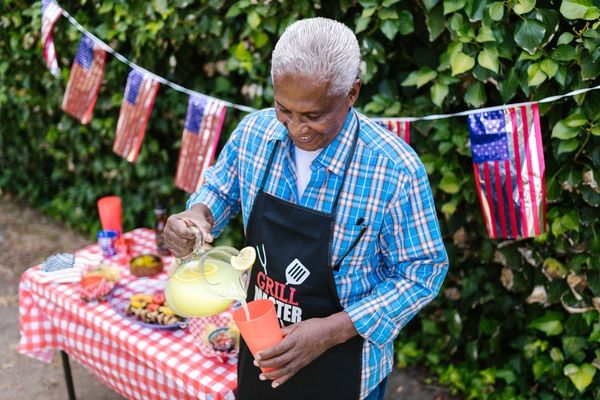 An older man wearing a “Grill Master” apron pours lemonade at an outdoor Labor Day cookout decorated with American flags and a red-checkered tablecloth.