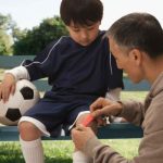 Parent applies a bandage to a young soccer player’s injured knee while the child sits on a park bench holding a soccer ball.