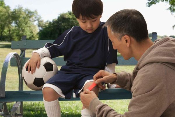 Parent applies a bandage to a young soccer player’s injured knee while the child sits on a park bench holding a soccer ball.