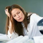 A tired woman sits at a desk, resting her head in one hand while looking at a coffee pot beside her laptop.