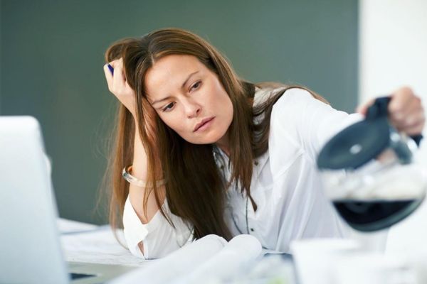 A tired woman sits at a desk, resting her head in one hand while looking at a coffee pot beside her laptop.