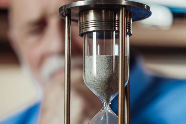 An hourglass in focus with an older man blurred in the background, representing the passage of time and aging.