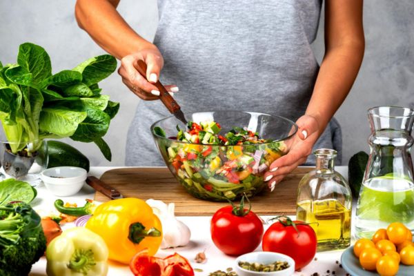 Person tossing a fresh vegetable salad surrounded by bell peppers, tomatoes, leafy greens, broccoli, and olive oil.